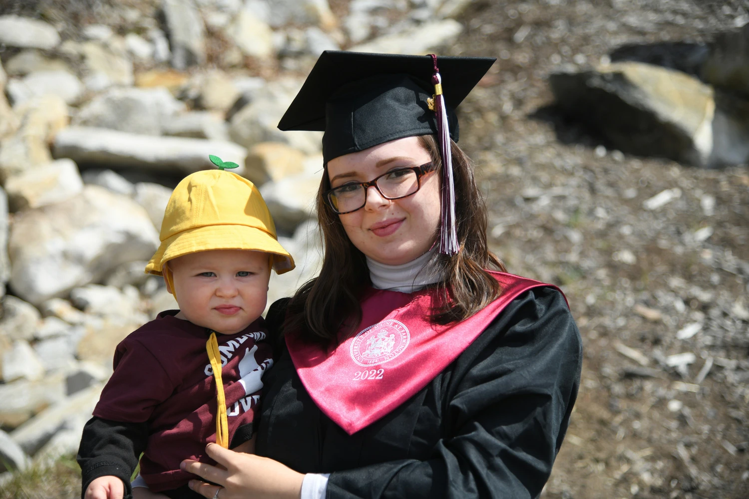 UMF Student in cap and gown hold baby.