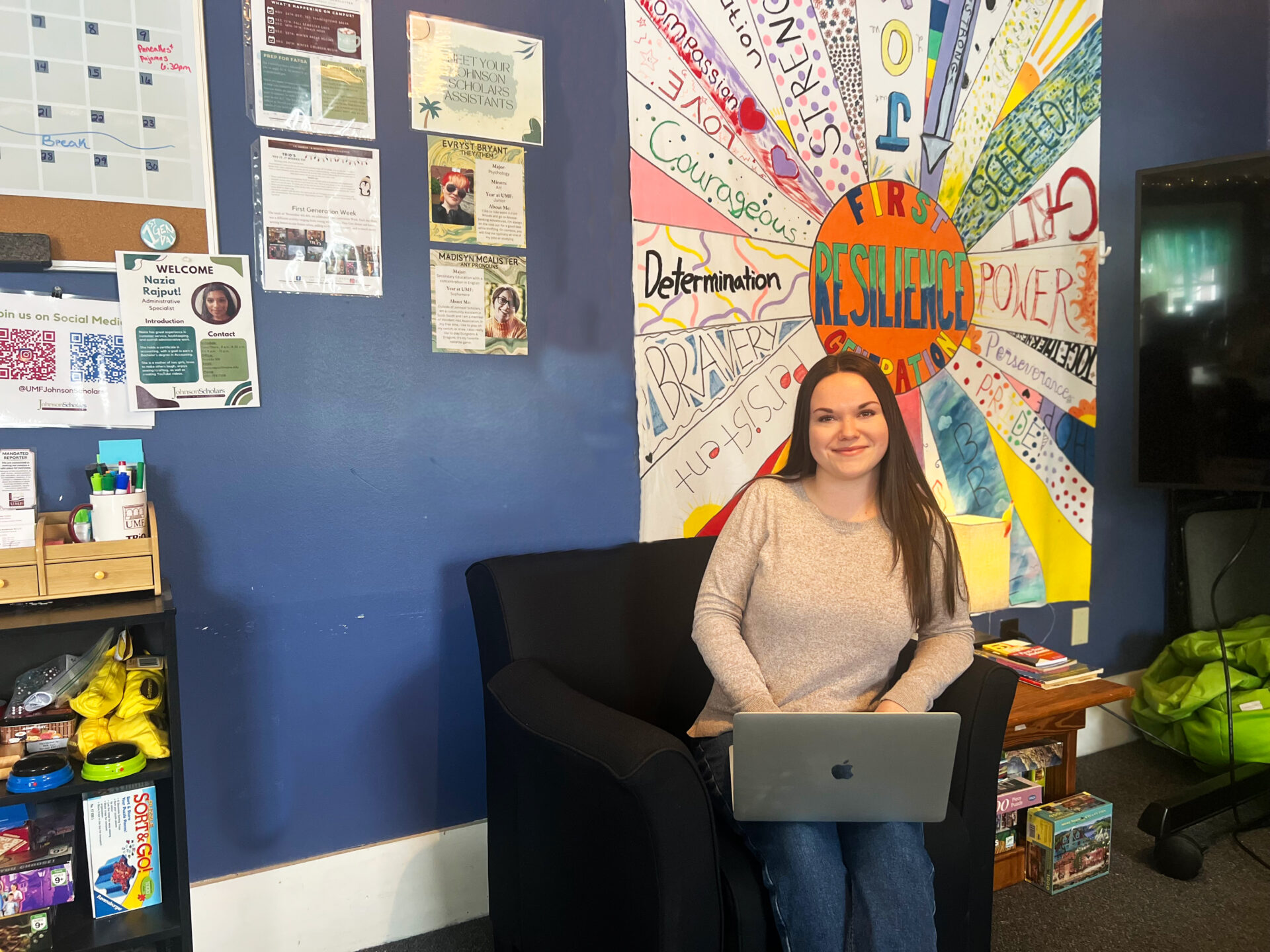 A student sitting in front of a bunch of posters and calendars, smiling at the camera as she types